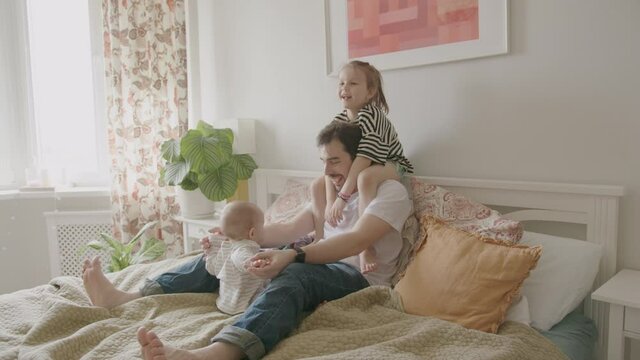 A Young Father and Two Baby Daughters, a Preschooler and an Infant, are Having Fun, Laughing and Jumping on the Bed. Cozy Home Interior.