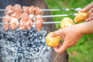 Hands of man prepares barbecue meat with potatoes on skewer by grill on fire outdoors. Concept of lifestyle rustic food preparation