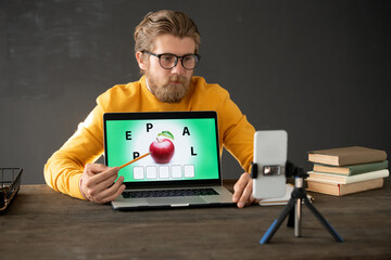 Young confident teacher with pencil pointing at red ripe apple on laptop display
