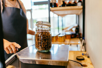 Female shop owner weighs glass jar with dark brown whole grains of pasta. Woman assistant in zero waste shop. Weighing dry goods in plastic free grocery store. Sustainable shopping at local business