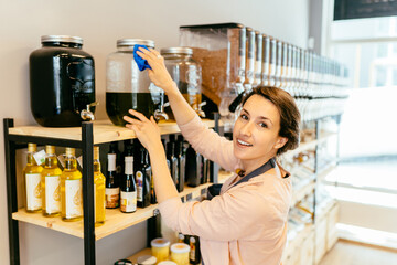 Owner woman with rag cleaning glass dispenser with metal tap with oil. Woman assistant in zero waste shop. Weighing dry goods in plastic free grocery store. Sustainable shopping at local business.