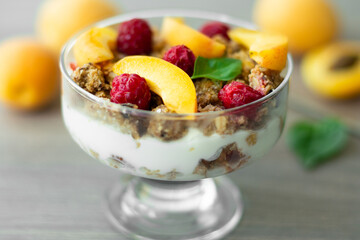 Healthy parfait with Greek yoghurt, homemade granola, slices of apricot and raspberries in a glass goblet. Grey wooden background with apricots and basil leaves