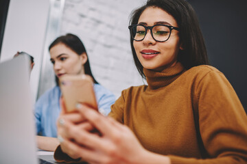 Positive young woman checking new feed in social networks via smartphone while doing homework in college, hipster girl sending text message in online chat with friend using cellphone while learning