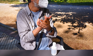 Fototapeta premium A man, sitting outdoors wearing a protective mask, cleans his hands with sanitizer lotion to eliminate germs and avoid infections. 