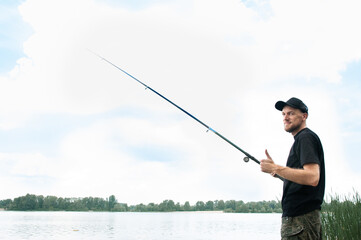 A fisherman in a black T-shirt, cap and camouflage shorts stands on the river bank with a classic fishing rod, free space for text