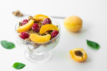 Healthy parfait with greek yoghurt, homemade granola, slices of apricot and raspberries in a glass goblet. White background with apricots and basil leaves