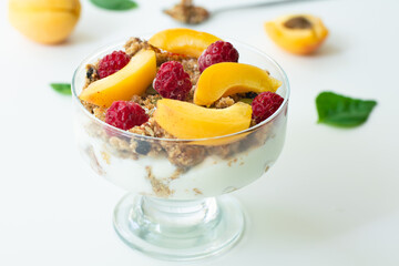 Healthy parfait with greek yoghurt, homemade granola, slices of apricot and raspberries in a glass goblet. White background with apricots and basil leaves