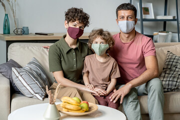 Young contemporary couple and their little son in masks sitting on couch