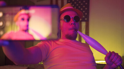 Blogger man smokes hookah and makes selfie photo by mobile phone on American flag background in dark room with pink neon lights.