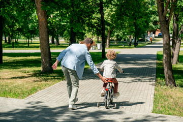 Back view of little boy in casualwear riding bicycle along road in public park