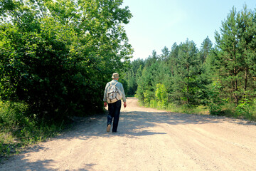 A man with a backpack is walking along a sandy road into the forest.