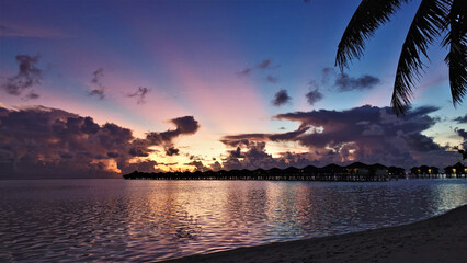 The sun disappeared beyond the horizon, leaving pink rays in the darkening sky. The first stars appeared above the picturesque clouds. In water villas, lights come on. Reflection in water. Maldives.