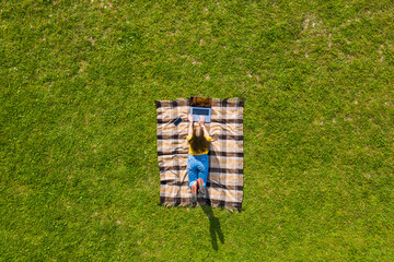 Drone view young woman working on laptop on green grass lying on plaid blanket