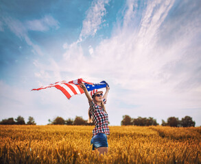 Beautiful young girl holding an American flag on the wind in a field of wheat. Summer landscape against the blue sky. Independence Day Fourth of July