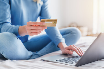 Close up of woman using credit card for payment on shopping online while sitting with laptop on bed in bedroom