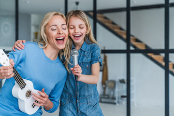 Beautiful mother and her cute little daughter with microphone singing together and playing ukulele