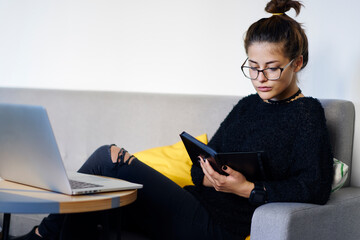 Concentrated student in optical eyeglasses reading interesting book sitting at laptop device on comfortable sofa.Pensive female manager checking plan of work process in notepad in cozy coffee shop