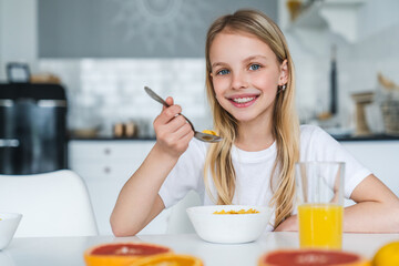 Cute little girl looking at camera while eating cereal balls with juice for her breakfast