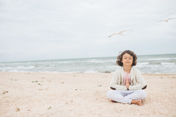 Young caucasian girl meditate at the beach, doing yoga lotus pose, child meditation outdoors