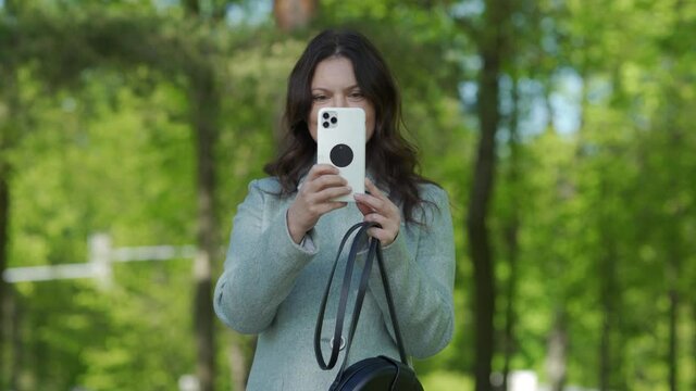 Adult woman smiling using camera on modern smartphone photographing in a park beautiful happy brunette woman holding mobile phone taking picture outdoors.