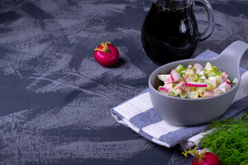 Okroshka soup with radish, sausage, cucumber, egg and potato in a ceramic bowl. Kvass in glass jug on the grey table. Russian cuisine meal
