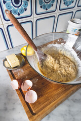 Glass bowl full of cake mixture, and ingredients on the side to make cake, sponge biscuits. On wooden board in kitchen