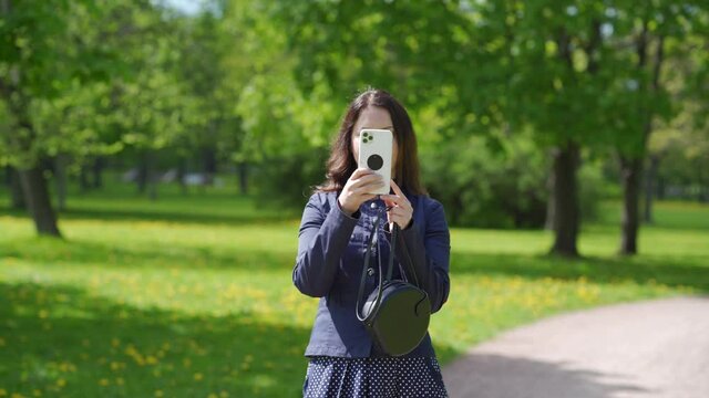Adult woman smiling using camera on modern smartphone photographing in a park beautiful happy brunette woman holding mobile phone taking picture outdoors.