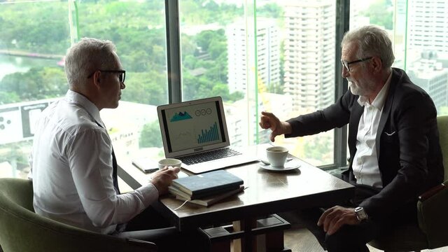 two senior businessman take a break drinking coffee and discussing business strategy on table with graph in laptop computer in coffee shop at modern restaurant on a tall building cityscape background