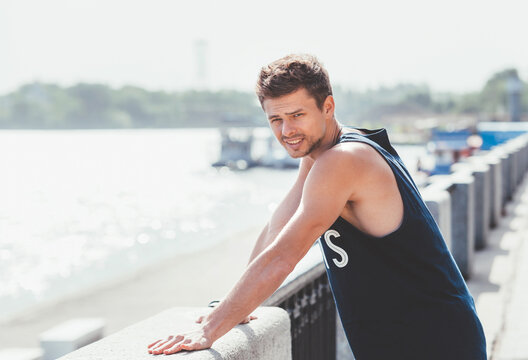 Active athlete muscular young man dressed sleeveless shirt resting after the jogging by the river promenade on the sunny day and enjoying the river view. Weekend sport activities concept image.