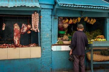 A colorful but dirty grocery store features  fresh meat hanging in the open windows along with...