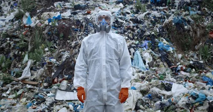 Male person in protective suit and respirator standing in front of huge trash hill. Man in protective clothing turning head and looking to camera. Concept of environmental pollution