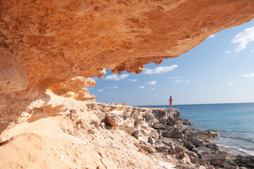 A young woman observes the horizon near a coastal cave. Formentera island, Mediterranean sea, Spain - obrazy, fototapety, plakaty