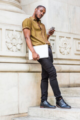 Young African American Man studying in New York, wearing green short sleeve shirt, black pants, leather shoes, carrying laptop computer, standing against white marble wall, texting on cell phone..