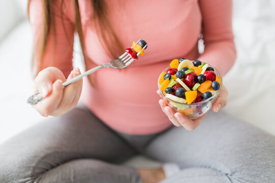 Close up of pregnant woman sitting on bed and eating fruit salad. Healthy meal concept.