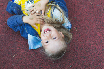Happy Caucasian girl with long blonde hair lies on the playground, hugs her hands to her chest and laughs with her mouth wide open.The milk tooth fell out.The child fell playing and laughs. From above