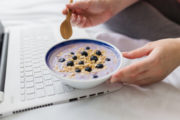 Young female eating granola and using laptop in bed, healthy breakfast and technology concept.