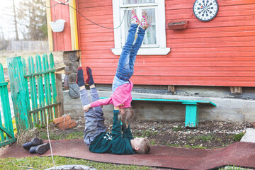 Caucasian children do acrobatic exercise on a track in a village near a wooden house. Boy lies on his back and supports a little girl who stands upside down. Training on self-isolation, a new reality.