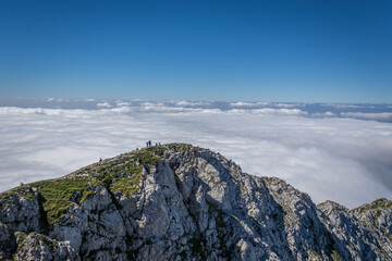 Aerial view of people on summit above low altitude fog. Inversion clouds covering large area. Alps mountain hiking. Hikers rest on rocky hilltop covered with grass on beautiful sunny day