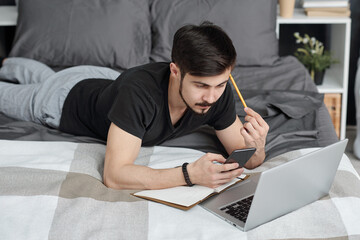 Brunette young man lying on bed with laptop and notepad and surfing social media on phone