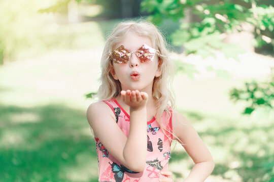 Young girl posing in fancy pink pentagonal shaped sunglasses outdoors. Cute adorable stylish Caucasian child with long blonde hair sending air kiss. Cool hipster kid having fun.