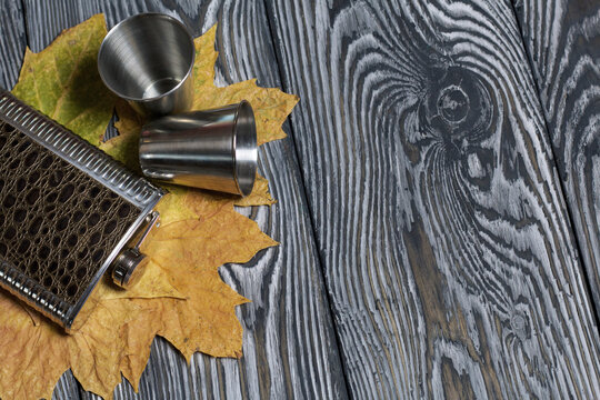 Flask for an alcoholic drink and metal glasses. Autumn maple leaves are yellow. On pine boards painted black and white.