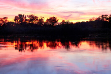 Colorful dramatic sunset sky over the river. Silhouettes of autumn trees on the river bank