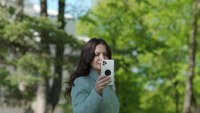 Beautiful adult woman taking picture by mobile phone in park brunette woman holding modern smartphone photographing on sunny spring day.