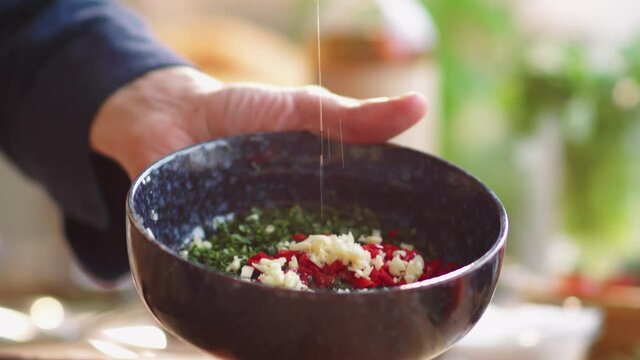 Close-up view of male cook pouring olive oil from glass bottle into bowl with fresh chopped vegetables while preparing salad