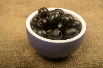 Black seedless olives in a ceramic bowl on a background of coarse-textured fabric. Close up.
