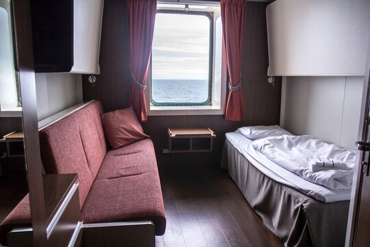 Interior of small but light cabin in passenger ship with window, bed and sofa.