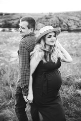 Pregnant girl with a tummy, awaiting delivery, in a red dress and a straw hat with her husband in a field and feather grass