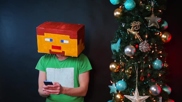 A man in a square mask on his head uses a smartphone. Nearby is a new Year tree. Black background.