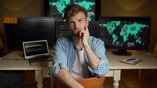 Portrait of thoughtful young man looking at camera with his hand on chin sitting in dark office with multiple computer monitors with programming code and global stock market data on screens