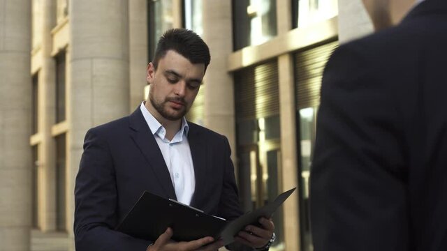 Young businessman in dark blue suit take folder with documents from his employee on the street near modern office building and talk with him about work.